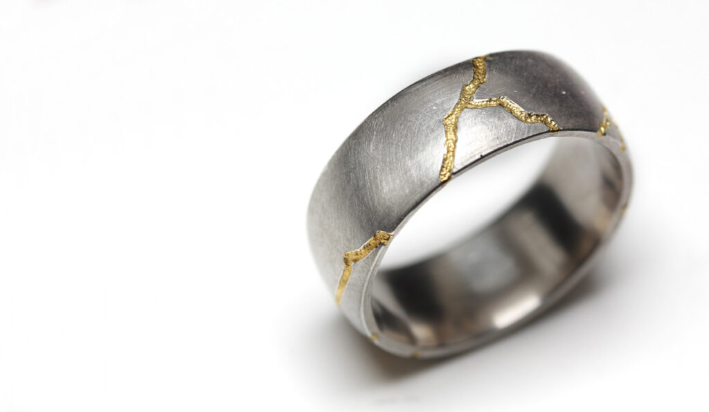 En ring i silver med inlägg av 24K guld. Tanken på att förhöja och omfamna ojämnheter och ”fel”, har styrt utformandet av denna ring. Mönstret är inspirerat av ”Kintsugi”, den japanska tekniken att reparera sprucken keramik. Istället för att dölja sprickorna framhävs de genom inlägg av guld eller silver. Erik Tidäng Smycken som bär på spår och lämnar avtryck